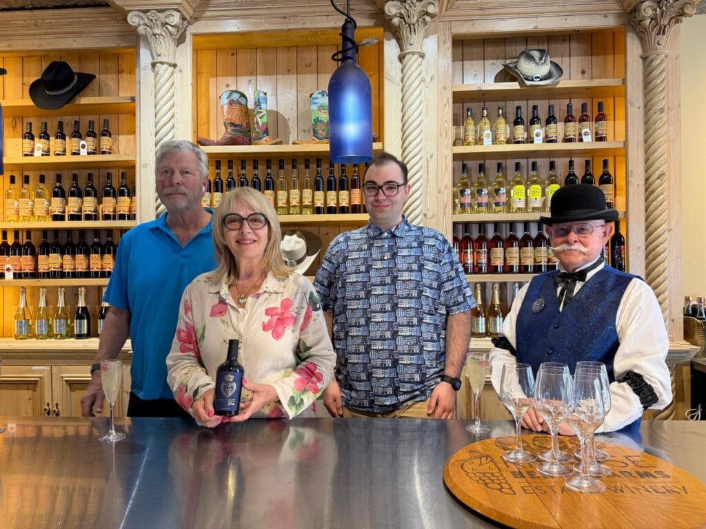 Al and Sandee Krause with two estate winery team members behind the tasting bar at Krause Berry Farms in Langley BC, with shelves of wine bottles and a staff member in a bowler hat and sparkly vest