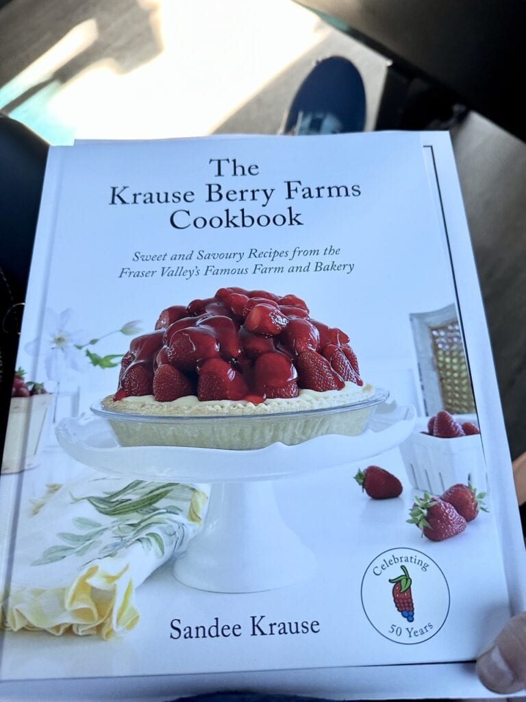 Hardcover cookbook with a fresh strawberry tart on the cover displayed on a white cake stand, held on a lap after visiting Krause Berry Farms in Langley BC