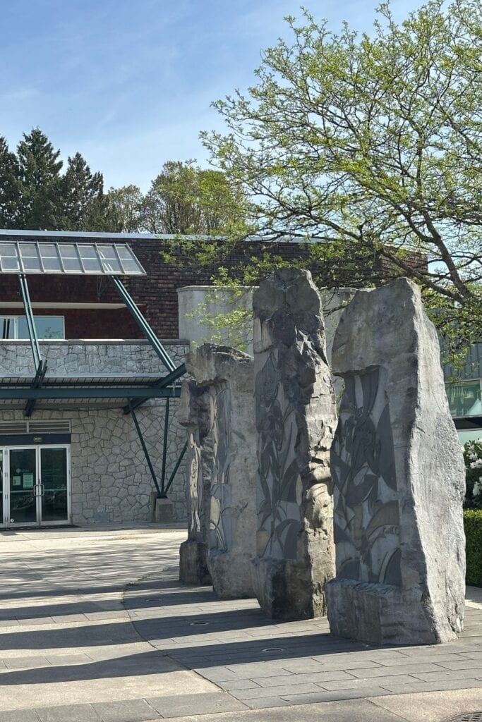 Large carved stone sculptures at the entrance of the Shadbolt Centre for the Arts in Burnaby, BC, with botanical carvings across the stone faces and the building's stone and glass exterior behind.