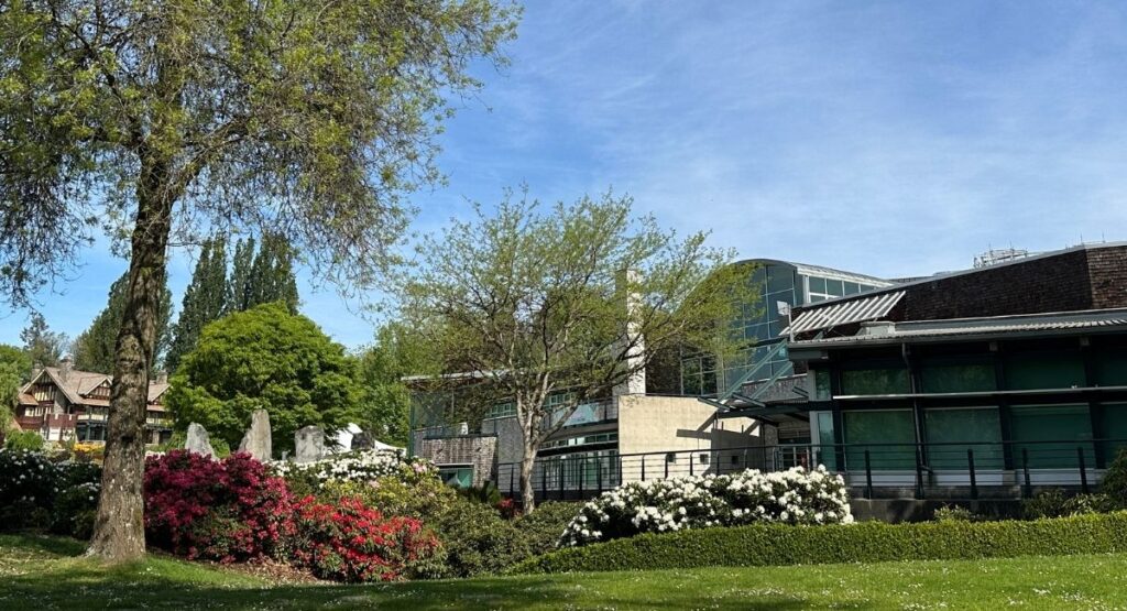 The Shadbolt Centre for the Arts in Burnaby, BC, a modern glass and steel building surrounded by blooming red and white rhododendron gardens, with the heritage Burnaby Art Gallery visible to the left and a blue sky above.