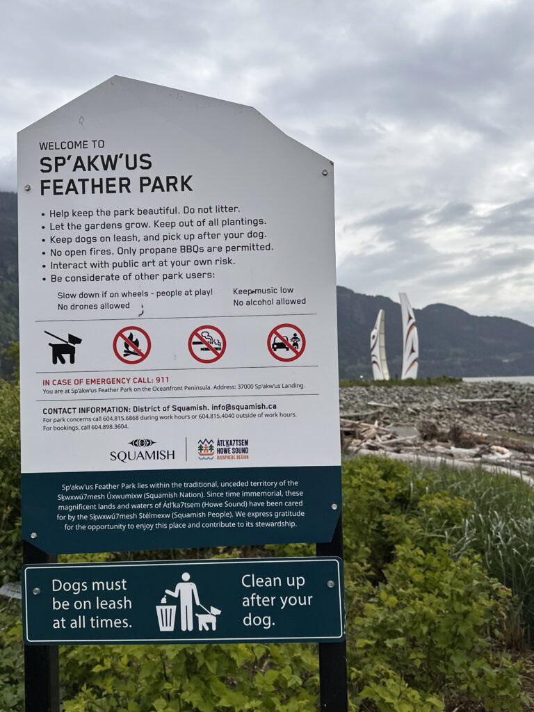 Park information sign at Sp'&aacute;kw'us Feather Park in Squamish, BC, with feather sculptures, shoreline plants, and mountain views for birdwatching.