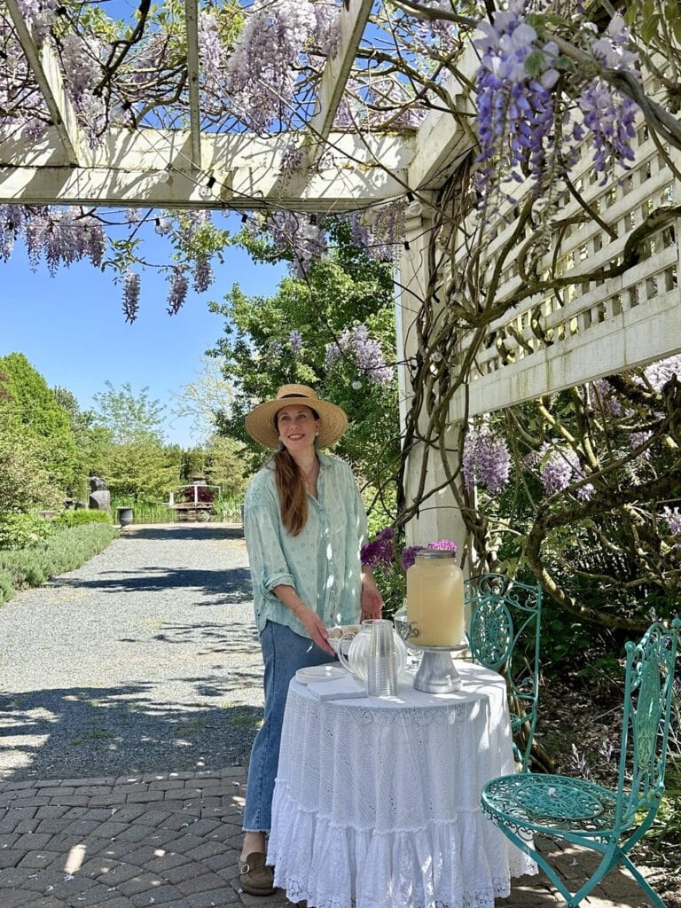 Woman in a straw hat under a white pergola covered in blooming purple wisteria at Tuscan Farm Gardens in Abbotsford, beside a table with lavender lemonade