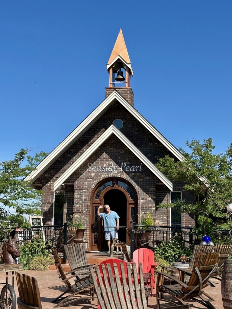 Chapel-style brick building with copper spire and bell at Seaside Pearl Winery in Abbotsford with a man waving from the arched doorway and wooden chairs on the brick patio