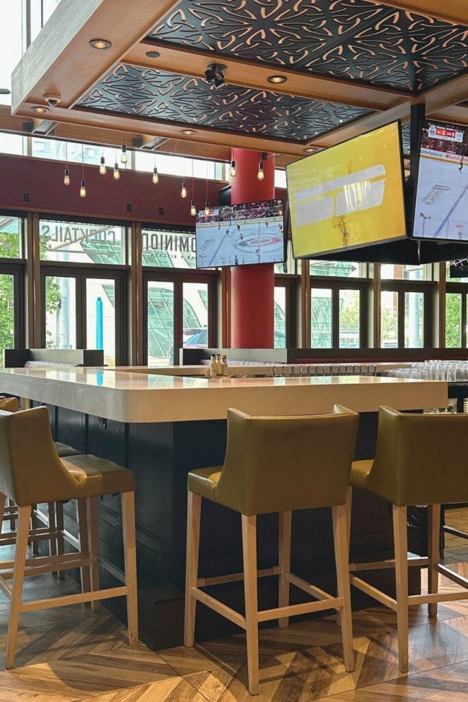 Interior bar area at Dominion Bar and Kitchen in Surrey City Centre BC with high stools, warm wood floors, patterned ceiling details and large screens showing a hockey game