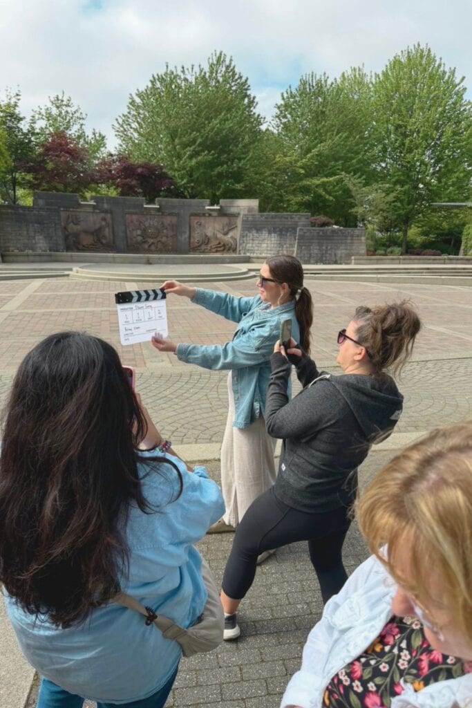A woman holding a film clapperboard in Holland Park Surrey BC while members of the Cineventure FAM tour group photograph and film the moment