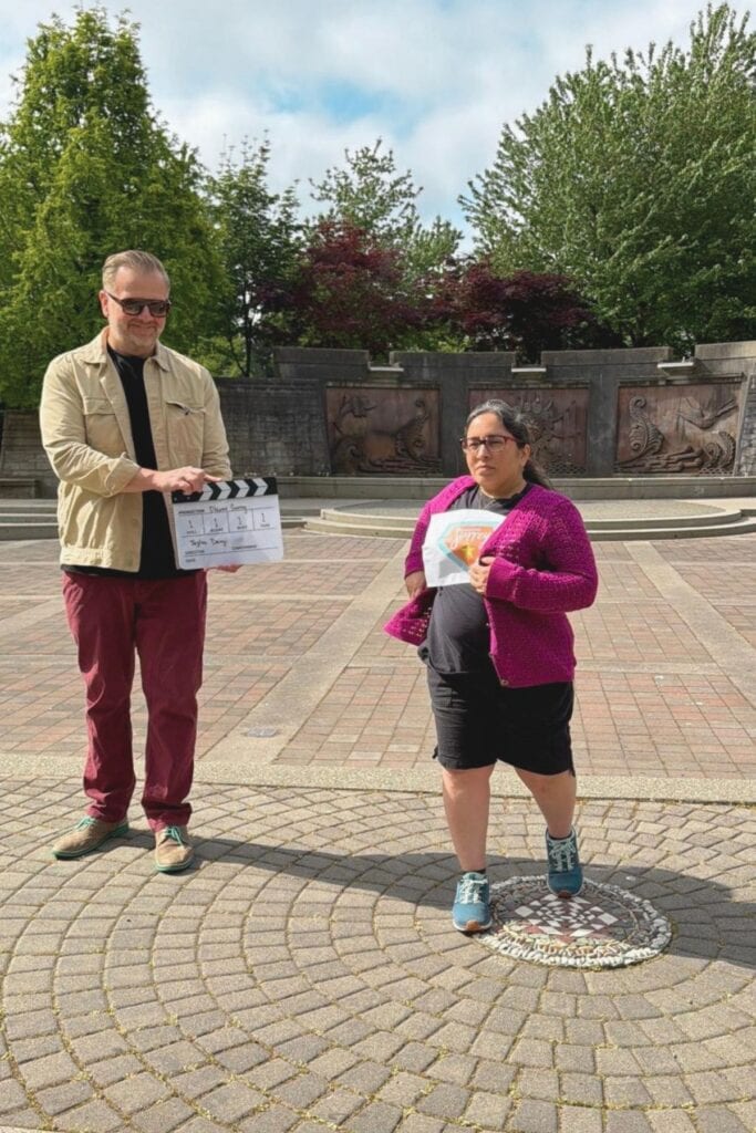 Tazim Damji standing in the Holland Park amphitheatre in Surrey BC holding a Discover Surrey bag while a man holds a film clapperboard beside her
