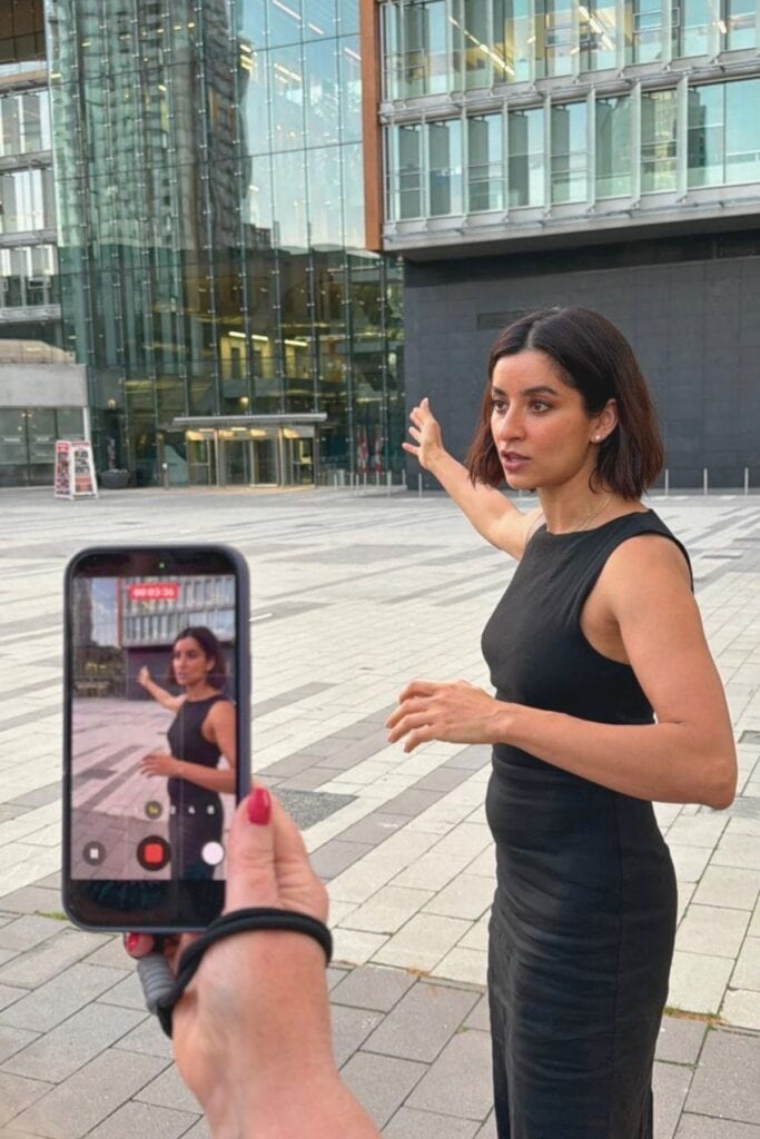 Supinder Wraich speaking and gesturing in Surrey Civic Plaza BC while someone films her on a smartphone, with the City Centre Library building in the background