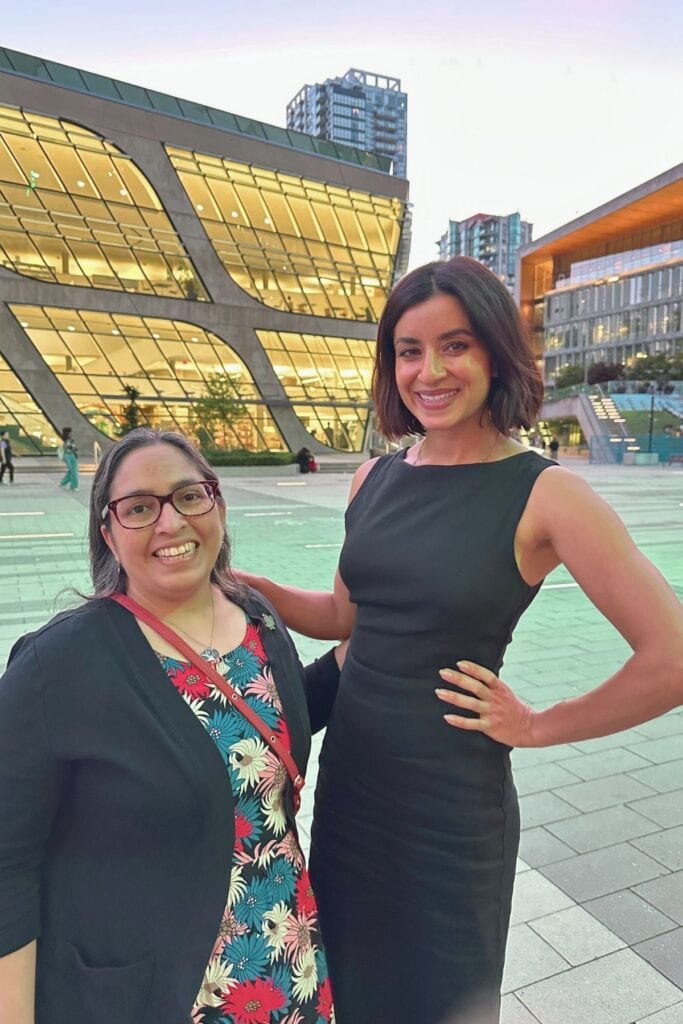 Tazim Damji and Supinder Wraich smiling together in front of the Surrey City Centre Library at dusk in Surrey BC