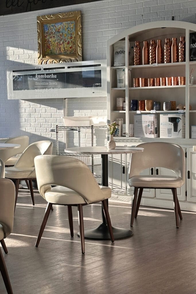 Sunlit café seating at Mezzo Market with cream upholstered chairs, round tables and shelves displaying copper bottles and cups against a white brick wall.