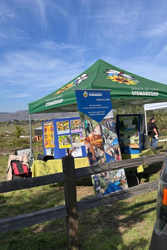 Okanagan Similkameen Stewardship booth at sncəcmałqtn Agricultural Park, with wildlife and habitat information displays beneath a green canopy.