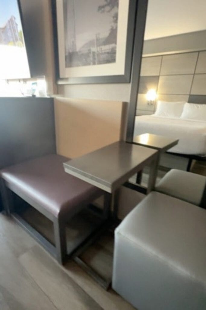 Guest room seating area at Best Western Premier Aberdeen Kamloops, featuring an upholstered bench, small rectangular table and cushioned chair. A large wall mirror reflects a bed with white bedding and a padded headboard.