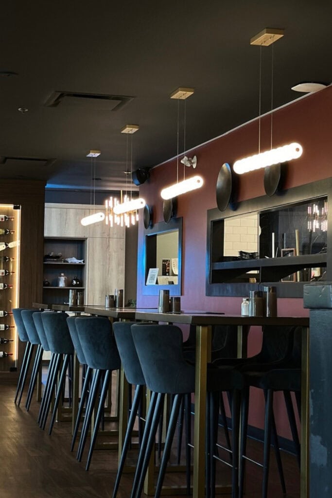 Interior of Heaven Sushi + Sake Bar at Best Western Premier Aberdeen Kamloops, featuring high-top tables, dark upholstered chairs and long pendant lights. A burgundy accent wall and modern decor frame the restaurant's seating area.