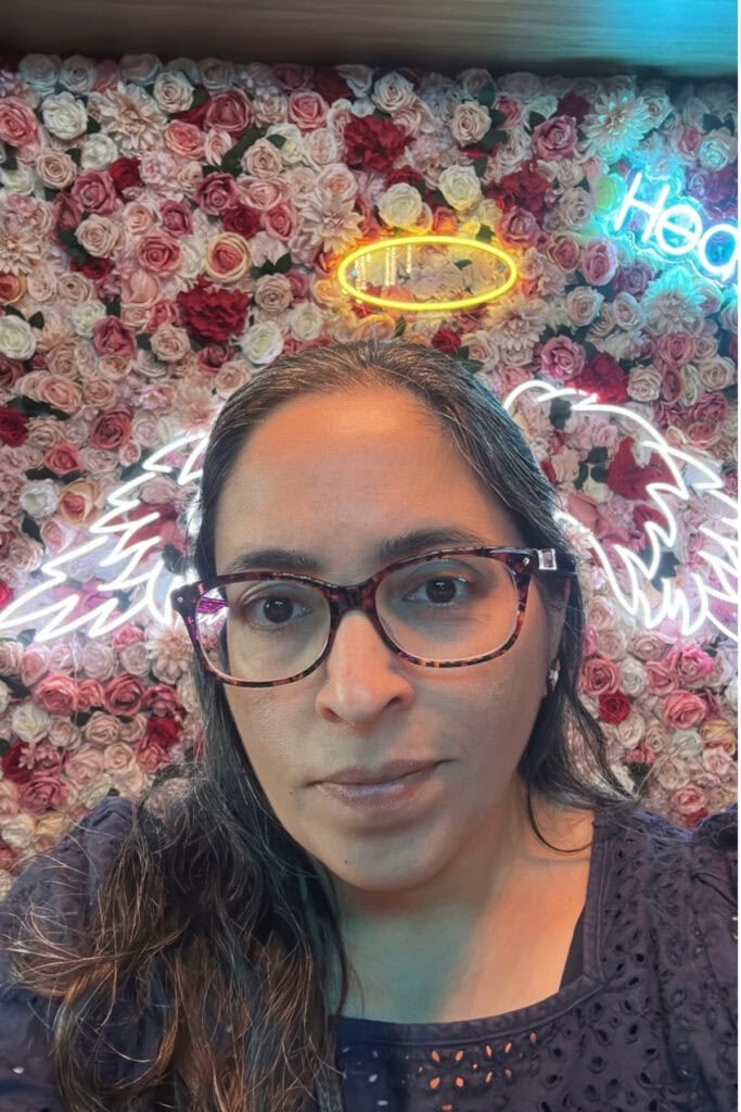 Selfie of a woman wearing glasses in front of a wall covered with pink, white and red artificial flowers at Heaven Sushi + Sake Bar in Best Western Premier Aberdeen Kamloops. Colourful neon lights and illuminated angel wings decorate the background.