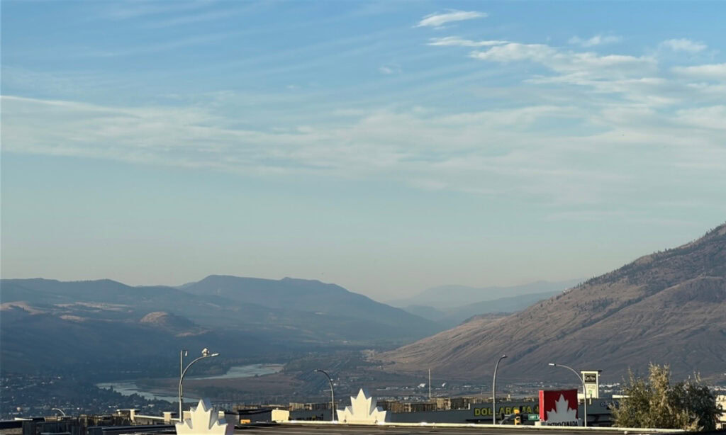 View across the Thompson Valley in Kamloops, BC, with dry hills, distant mountains and the Thompson River winding through the landscape. Commercial buildings and Canadian flag decorations are visible in the foreground near Best Western Premier Aberdeen
