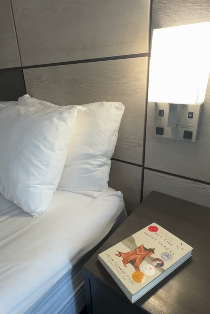 Close-up of a guest room bed with white pillows, a wood-panelled headboard and a wall-mounted reading light at Best Western Premier Aberdeen Kamloops. A copy of All the Quiet Places by Brian Thomas Isaac rests on the bedside table.