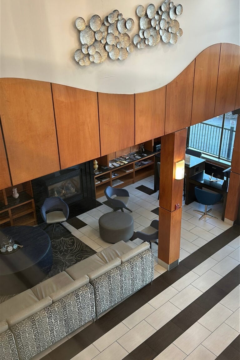 View overlooking the spacious lobby of Best Western Premier Aberdeen Kamloops, featuring curved wood panelling, decorative wall sculptures, lounge seating and a fireplace. The reception desk and business centre are visible below.