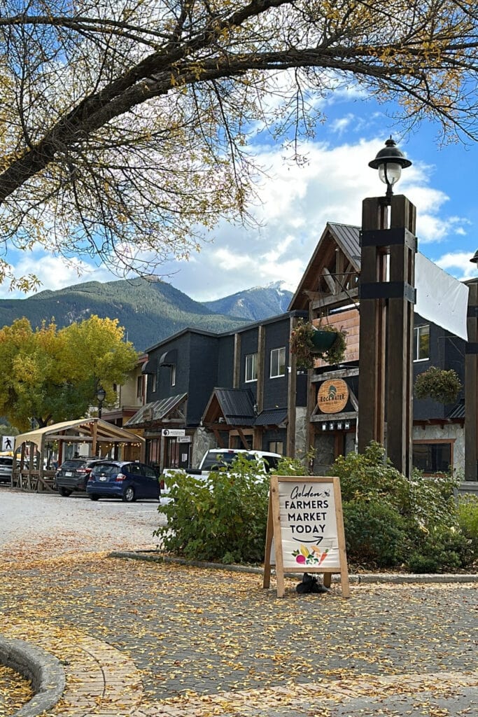 Golden Farmers' Market sign on a sidewalk covered in yellow autumn leaves, surrounded by downtown Golden shops and mountains.