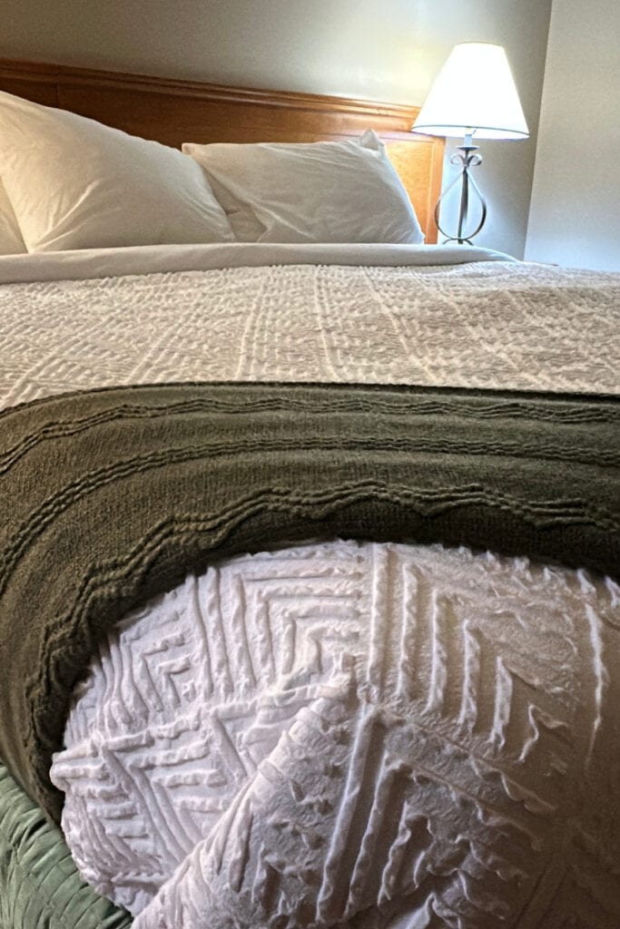 Bed with white textured bedding and a dark green throw in a guest suite at Palliser Lodge, Kicking Horse Mountain Resort near Golden, BC.