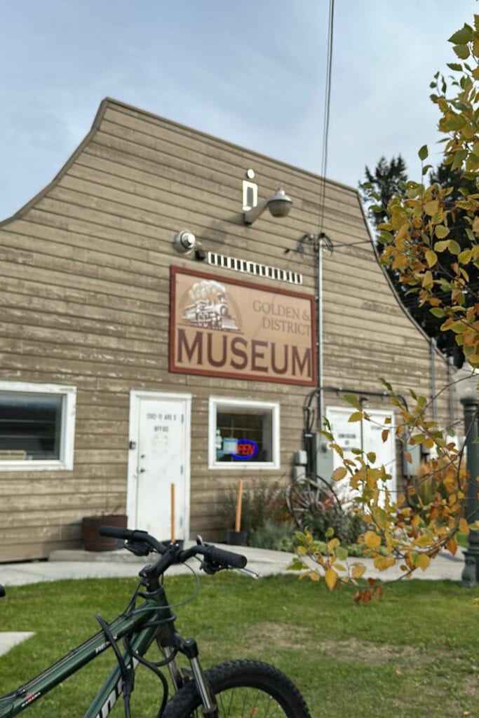 Exterior of the Golden & District Museum in Golden, BC, with a bicycle parked outside and yellow autumn leaves beside the historic-looking building.
