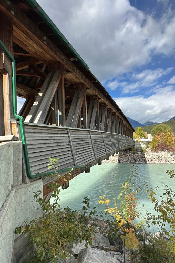 Side view of the timber-framed Kicking Horse Pedestrian Bridge crossing the turquoise Kicking Horse River in Golden, BC, surrounded by autumn foliage.