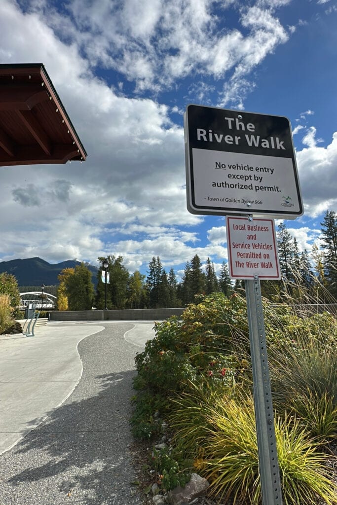 The River Walk sign beside a paved riverside pathway in Golden, BC, with autumn plants and the Kicking Horse Pedestrian Bridge in the distance.