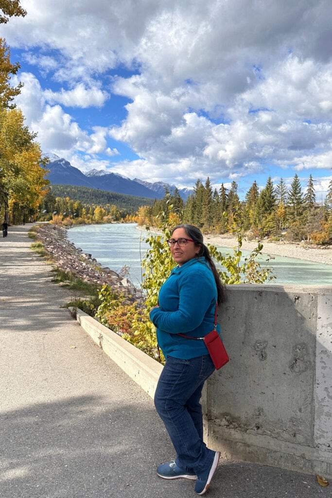 Travel writer beside the turquoise Kicking Horse River in Golden, BC, with yellow fall foliage and snow-dusted Canadian Rockies mountains.