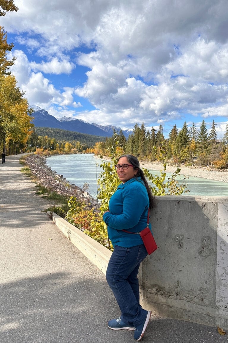 Travel writer beside the turquoise Kicking Horse River in Golden, BC, with yellow fall foliage and snow-dusted Canadian Rockies mountains.