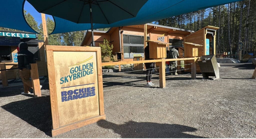 The Golden Skybridge Rockies Rangers sign stands near the ticket booths, gift shop and wooden entrance buildings beneath large blue shade canopies. This is part of the main arrival area where visitors enter the attraction.
