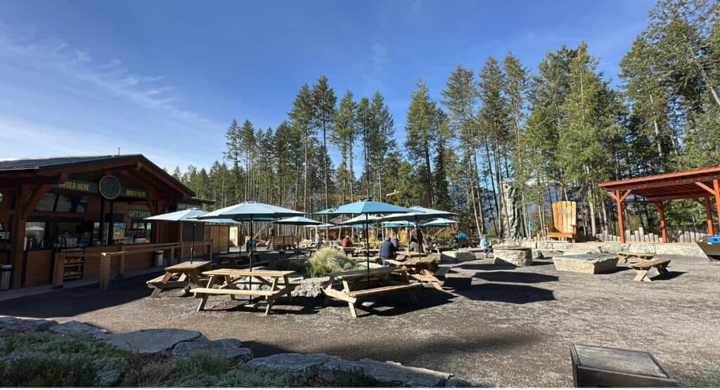 Wooden picnic tables and blue umbrellas fill a spacious outdoor seating area at Golden Skybridge, with a food counter and forest trees surrounding the plaza. The Village offers a place for visitors to sit, eat and take a break between activities.