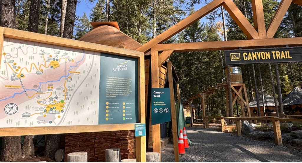 A large illustrated trail map stands beside the timber-framed Canyon Trail entrance at Golden Skybridge in Golden, BC. The gravel pathway leads into the forest, with directional signs and visitor information beside the entrance.