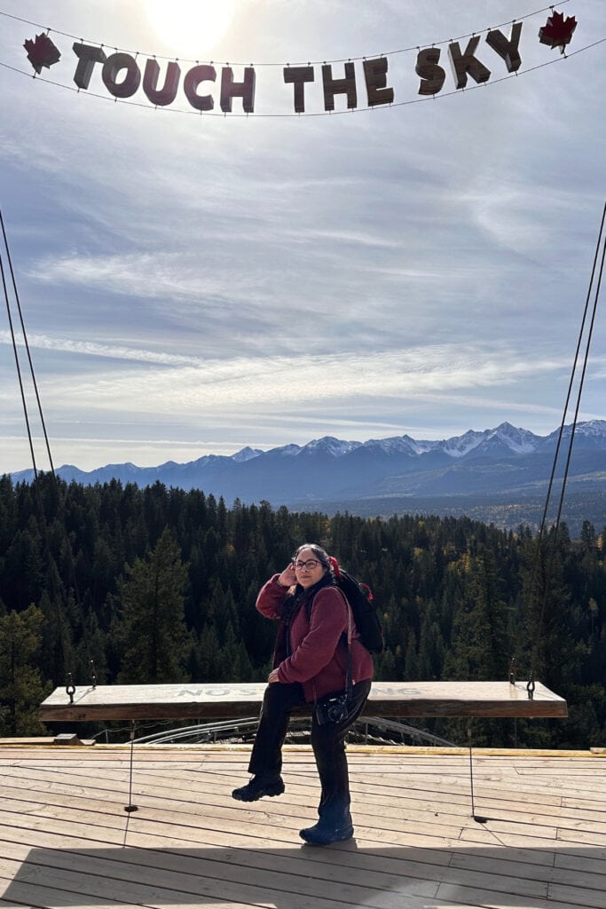 Canadian travel writer Tazim Damji poses on a large wooden swing beneath the "Touch the Sky" sign at Golden Skybridge in Golden, BC. Forested hills, the Railrider Mountain Coaster and snow-capped mountains create a scenic backdrop for this popular photo spot.