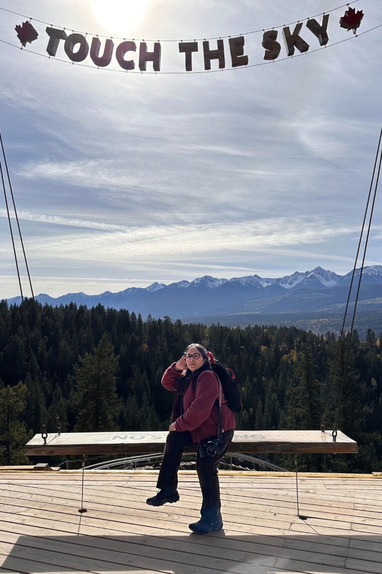 Canadian travel writer Tazim Damji poses on a large wooden swing beneath the "Touch the Sky" sign at Golden Skybridge in Golden, BC. Forested hills, the Railrider Mountain Coaster and snow-capped mountains create a scenic backdrop for this popular photo spot.