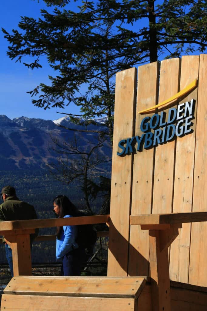 A large wooden Golden Skybridge sign stands beside a mountain viewpoint in Golden, British Columbia, with forested slopes and snow-covered peaks in the background. Visitors can stop at this lookout to enjoy views of the surrounding landscape.