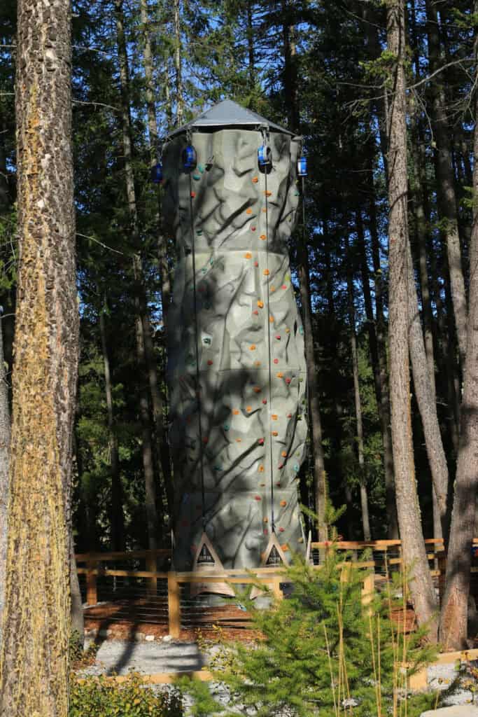 A tall, grey outdoor climbing tower with colourful handholds and hanging safety ropes stands among evergreen trees at Golden Skybridge in BC. The climbing wall offers an additional adventure activity within the forested attraction.
