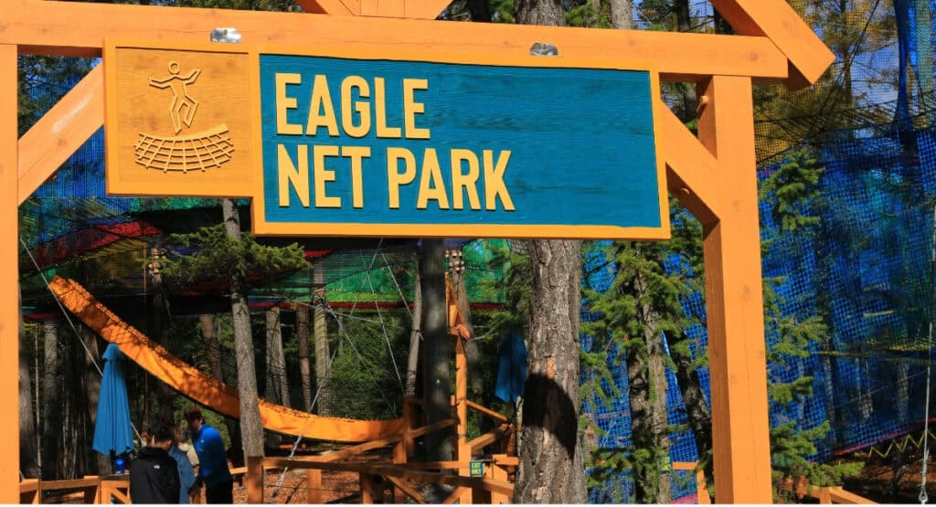 A large wooden Eagle Net Park entrance sign marks the elevated net adventure attraction at Golden Skybridge in Golden, BC. Colourful suspended netting, orange connecting sections and forest trees are visible behind the sign.