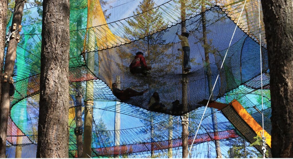 Visitors climb and lounge inside suspended net platforms at Golden Skybridge's Eagle Net Park, surrounded by tall evergreen trees. Blue, yellow, green and orange netting connects the different elevated play areas.