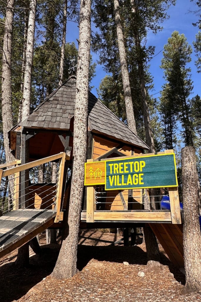 A wooden Treetop Village sign stands beside an elevated timber playhouse connected to a raised walkway at Golden Skybridge in Golden, BC. The forest play area features wooden structures built among the trees.