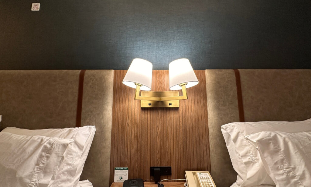 Two white pillows resting against upholstered headboards with a double wall-mounted reading lamp between them at Holiday Inn & Suites Kamloops, BC. A bedside telephone and electrical outlets are positioned below the light.