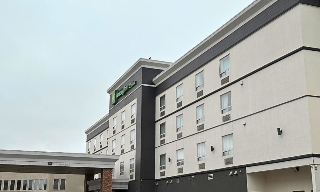 Exterior of Holiday Inn & Suites Kamloops, BC, showing the multi-storey hotel building with grey and white walls, rows of guest room windows and the green Holiday Inn sign. A covered entrance extends from the front of the property.