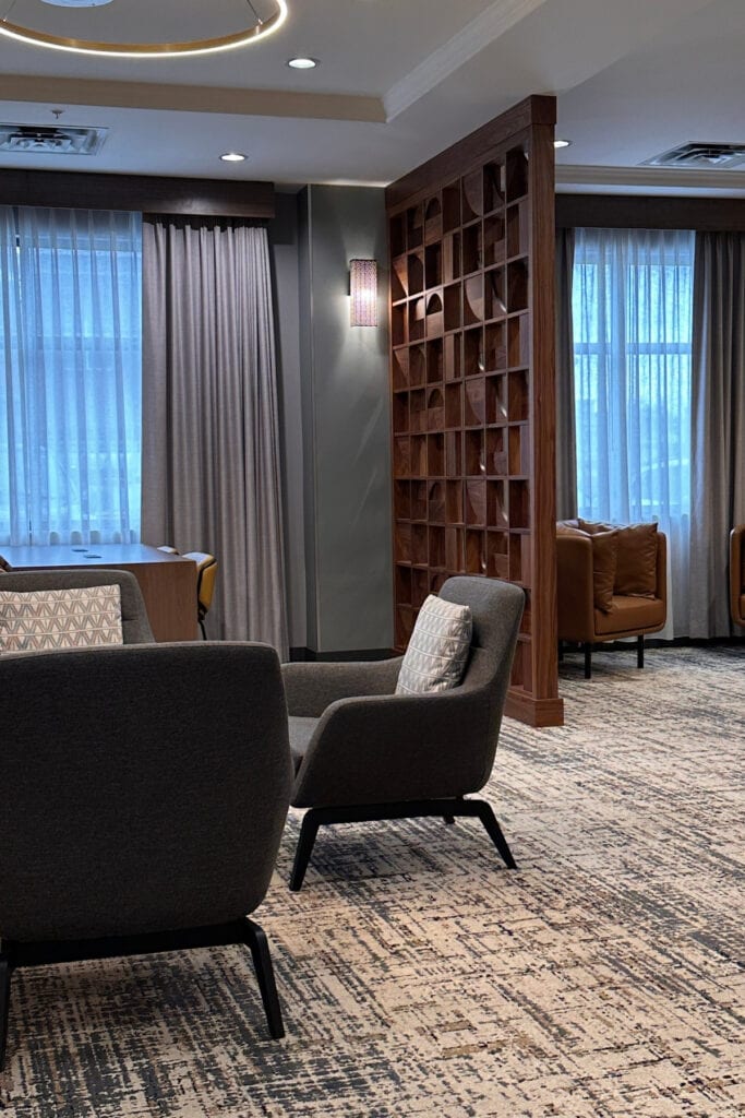 Grey upholstered armchairs, a brown leather chair and a geometric wooden room divider in the lounge at Holiday Inn & Suites Kamloops, BC. Large windows with sheer curtains provide natural light to the seating area.