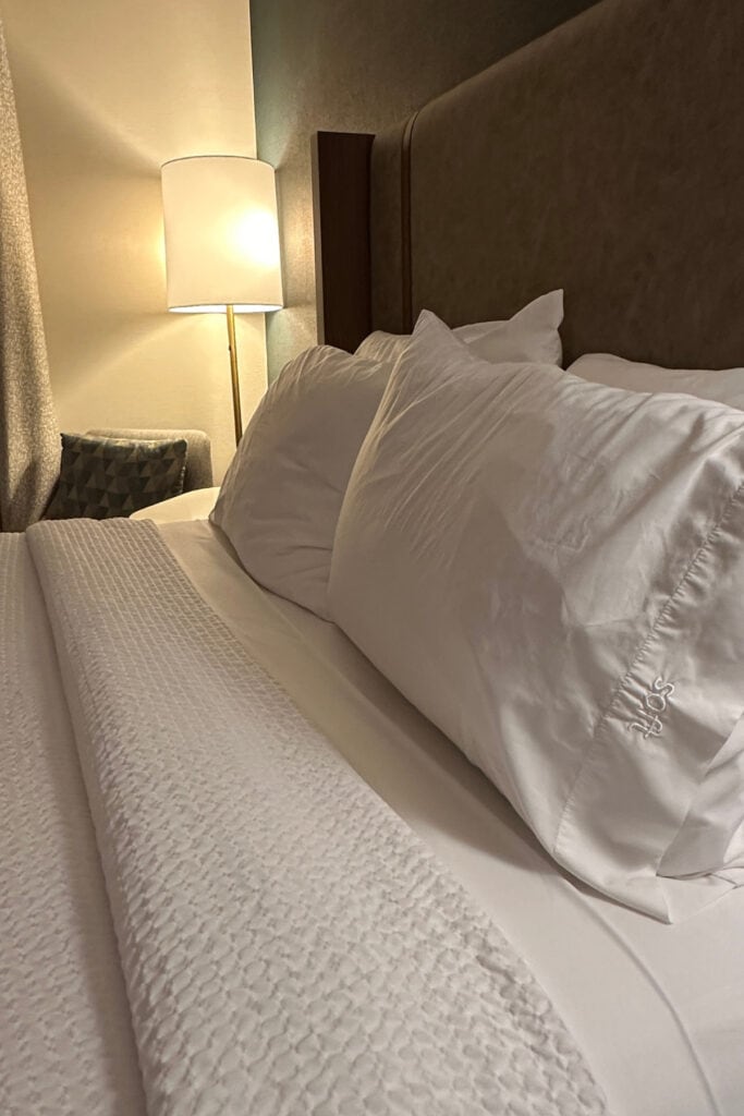 Close-up of a hotel bed with white pillows, a textured white duvet and a tall upholstered headboard at Holiday Inn & Suites Kamloops, BC. A floor lamp and cushioned chair sit beside the bed.