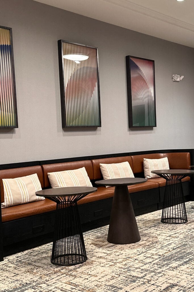 Brown leather bench seating with striped cushions, small black tables and framed abstract artwork in a common area at Holiday Inn & Suites Kamloops, BC. The seating arrangement sits against a grey wall above patterned carpeting.