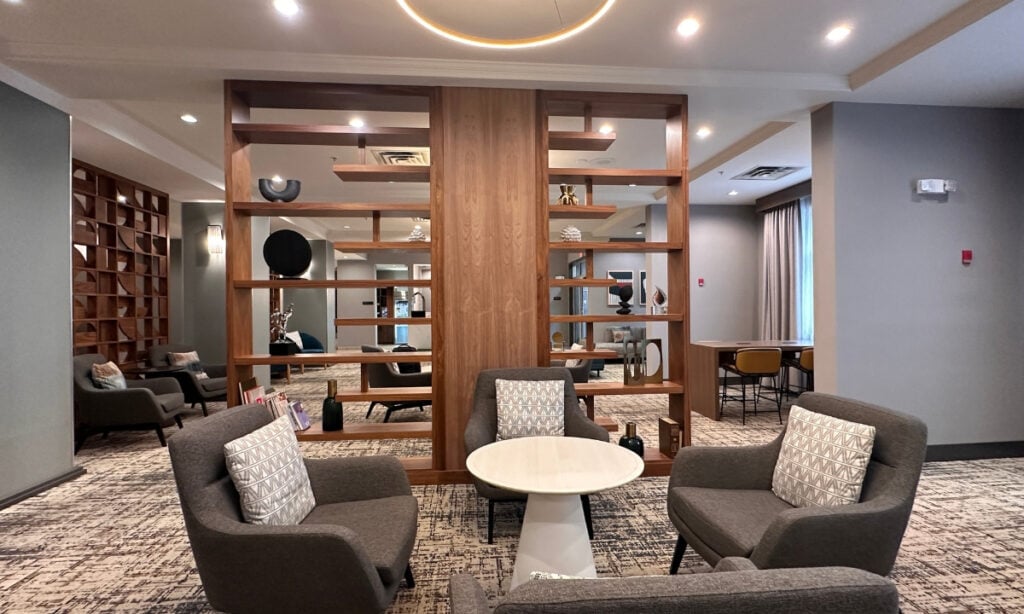 Grey upholstered armchairs arranged around a white circular table in the lounge at Holiday Inn & Suites Kamloops. Open wooden shelving divides the seating areas, with additional chairs and a worktable visible behind it.
