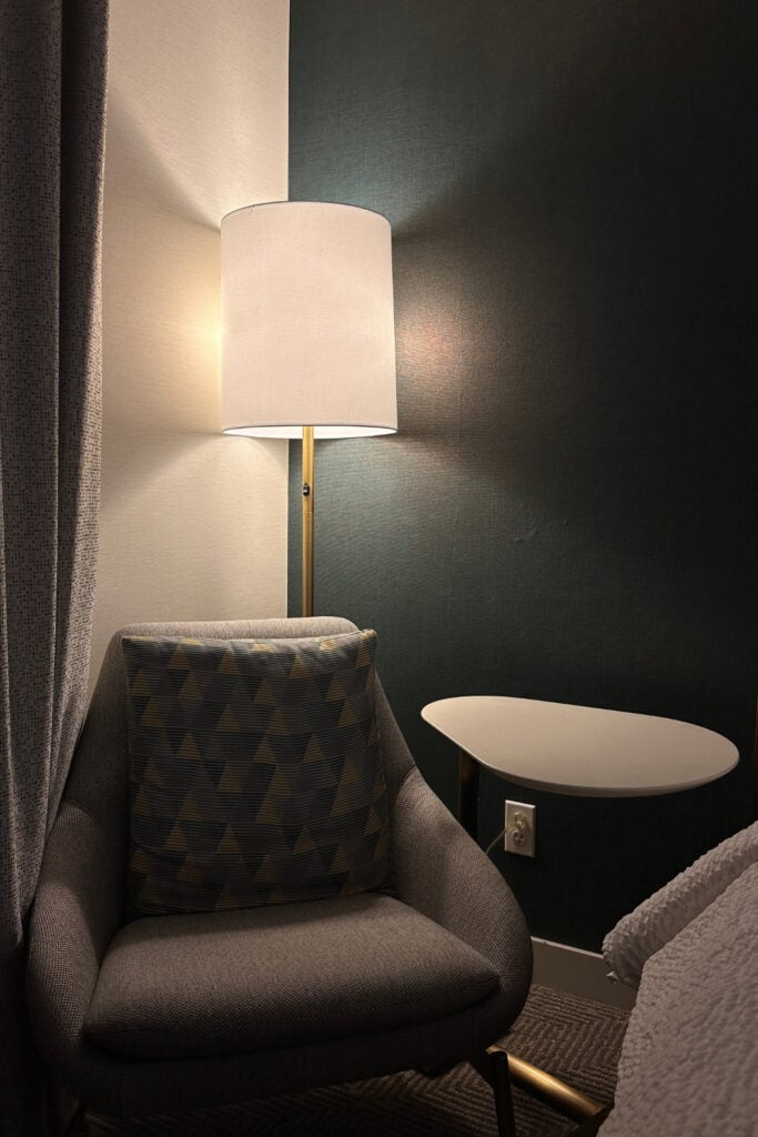 Grey upholstered armchair with a geometric-patterned cushion beside a tall floor lamp and small wall-mounted table at Holiday Inn & Suites Kamloops, BC. Dark accent walls and curtains surround the guest room seating area.