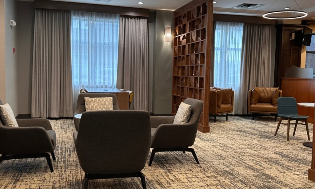 Wide view of the Holiday Inn & Suites Kamloops lounge with grey armchairs, brown leather chairs, large curtained windows and a geometric wooden partition. Patterned carpeting covers the floor throughout the seating area.