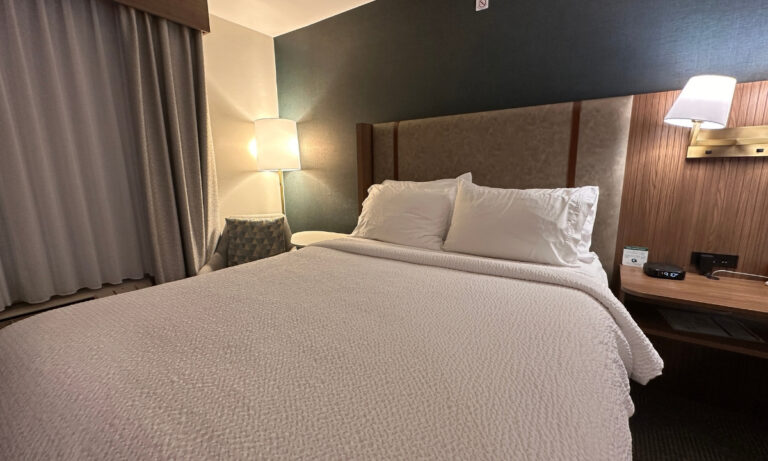 Guest room at Holiday Inn & Suites Kamloops, BC, featuring a large bed with white bedding, an upholstered headboard and wall-mounted reading light. A cushioned chair, floor lamp and bedside table complete the room.