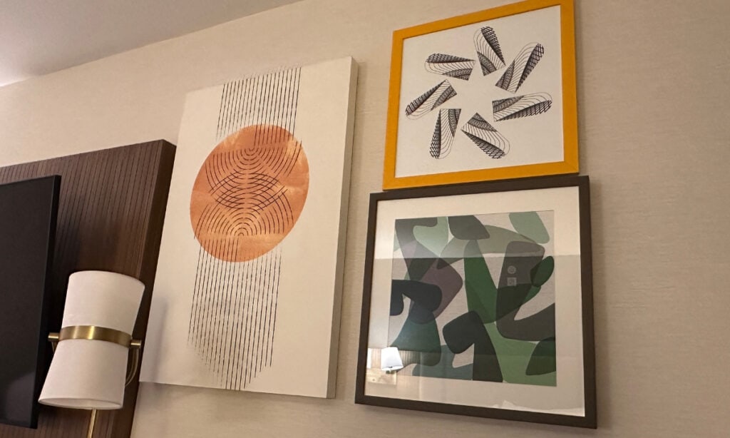 Three framed abstract artworks featuring orange, black, green and geometric patterns displayed on a guest room wall at Holiday Inn & Suites Kamloops, BC. A modern wall-mounted light fixture is visible beside the artwork.