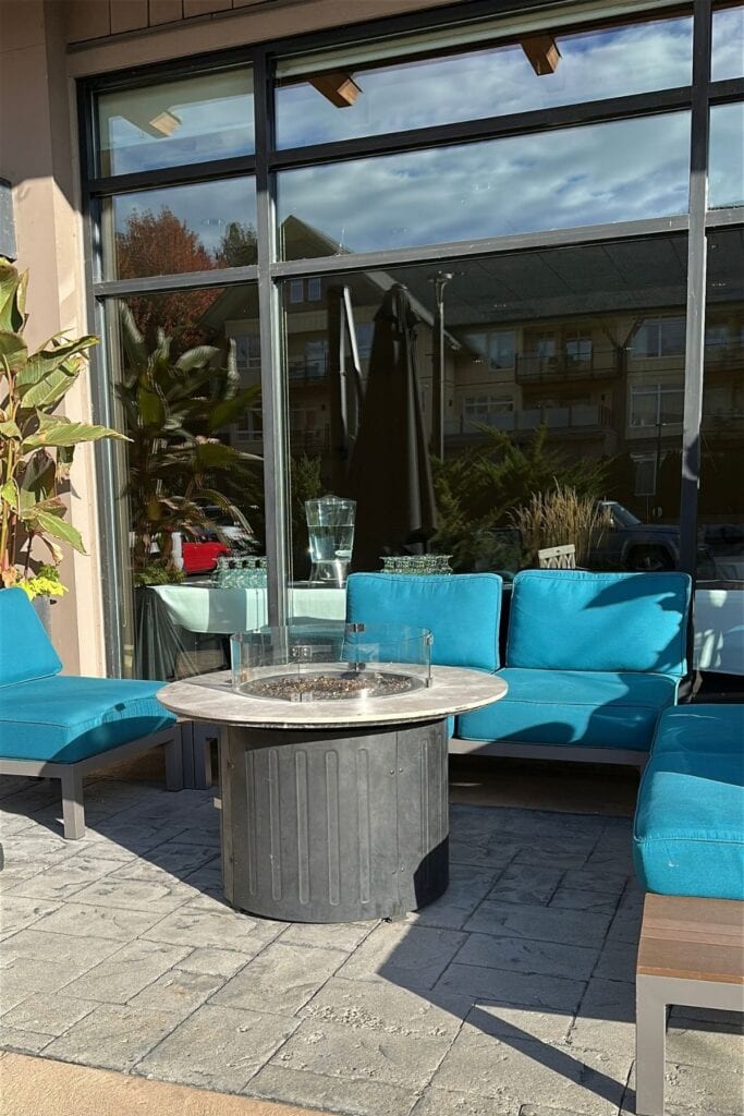 Outdoor seating area at Summerland Waterfront Resort and Spa featuring bright turquoise cushioned sofas surrounding a round gas fire pit with a clear glass wind guard. Large glass windows reflect neighbouring buildings and trees behind the sunny stone patio.