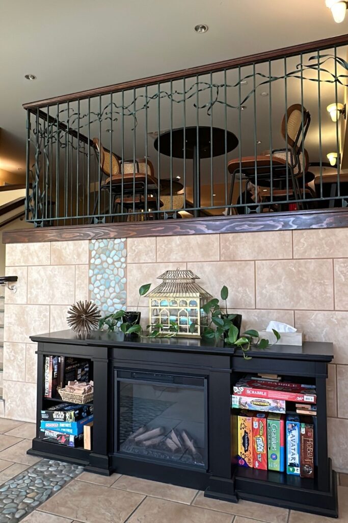 A black electric fireplace surrounded by shelves of board games including Scrabble, Cranium, Avatar and The Hobbit inside Summerland Waterfront Resort and Spa. A decorative lantern and green plants sit above the fireplace with an upper level seating area visible behind a metal railing.