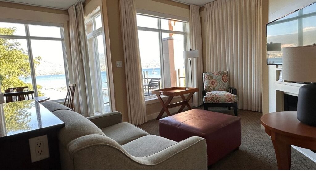 Bright living room at Summerland Waterfront Resort and Spa featuring a grey sofa, burgundy leather ottoman, floral armchair and large windows overlooking the lake. Floor to ceiling cream curtains frame the waterfront views and a television hangs above the fireplace.